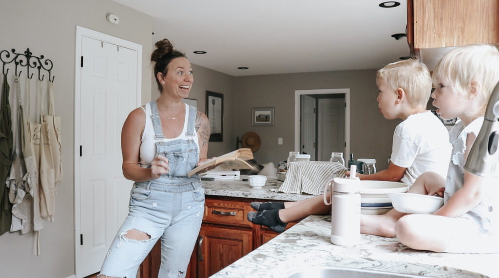 Christian mom wearing a white tank top and overalls reads a book to her two children that are sitting on the kitchen counter